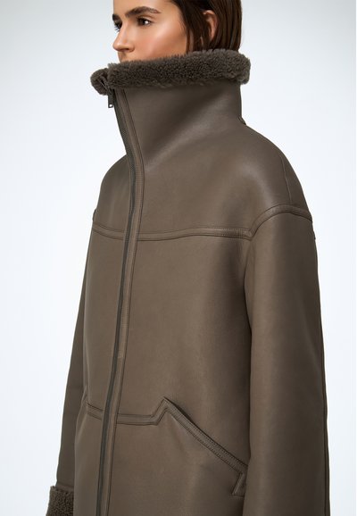 Chaqueta de cuero marrón con cuello alto y forro de borrego. Cuenta con una cremallera frontal y bolsillos laterales en ángulo, mostrando una textura suave.