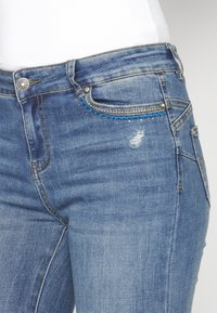 Gros plan sur un jean en denim bleu avec des surpiqûres décoratives et une petite zone usée sur la poche avant.