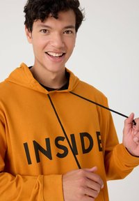 Sudadera con capucha naranja de algodón, con el texto negro "INSIDE" en la parte frontal, cuenta con un cordón negro y puños acanalados.