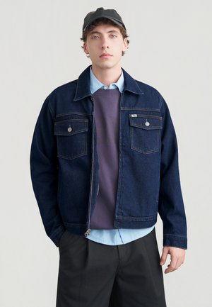 Tmavomodrá rifľová bunda s dlhými rukávmi, dvoma náprsníkovými vreckami, strieborným kovaním, vrstvená cez fialovú mikinu a svetlomodrú košeľu.