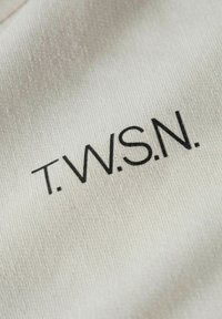 Tessuto beige con una superficie liscia, caratterizzato dal testo stampato in nero "T.W.S.N." in un moderno font sans-serif, centrato sulla superficie.