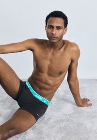 Boxer-briefs noirs Nike avec une ceinture élastique bleu vif, texture lisse et coupe ajustée, présentés sur un fond gris.