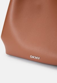 Torba DKNY wykonana z teksturowanego brązowego materiału syntetycznego. Charakteryzuje się srebrnym akcentem logo na dole. Gładki, stukturalny design o kanciastej formie.