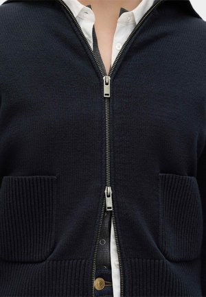 Cardigan blu navy con zip e due tasche frontali, indossato sopra una camicia bianca con bottoni e jeans scuri.