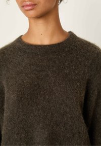 Pull en laine bouclée marron foncé, avec un col rond et des manches longues. Texture douce, design minimal, sans motifs ni accessoires visibles.