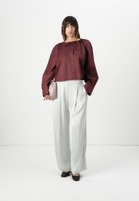 Maroon cropped button-up shirt with bow tie detail, paired with light green pleated trousers. Model holds a small pink handbag and wears black shoes.