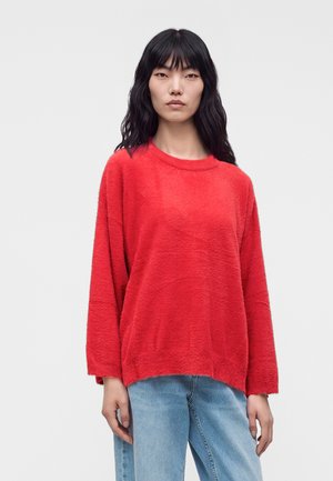 Femme aux longs cheveux foncés portant un pull rouge ample et un jean bleu clair, debout devant un fond clair uni.