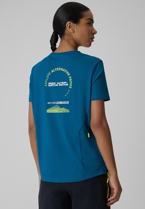 Kobieta z ciemnymi włosami w warkoczu, ubrana w niebieski T-shirt z żółtym i białym tekstem na plecach o eksploracji alternatywnych ścieżek.