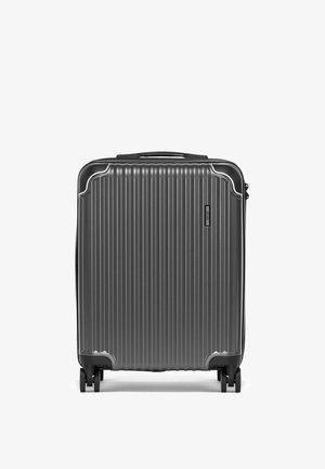 Ciężka walizka o twardej powłoce w ciemnoszarym kolorze z pionowymi żebrowaniami, górnym uchwytem i czterema obrotowymi kółkami, stojąca pionowo na białym tle.