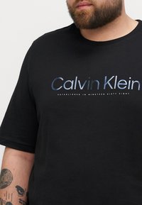 Svart bomull T-skjorte med et reflekterende blått og sølvfarget "Calvin Klein"-logo med teksten "ETABLERES I NITTENDE SEKSTI ÅTTE."