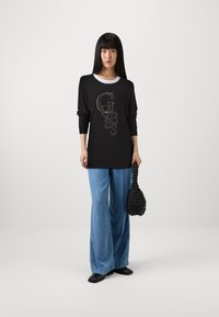 Top nero a maniche lunghe con stampa del logo, abbinato a jeans a gamba larga azzurri. Porta una borsa nera testurizzata e scarpe nere basse. Sfondo semplice.