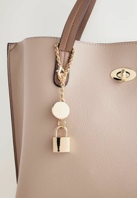 Sac cabas en cuir synthétique beige avec poignées marron, doté d'une chaîne en or et d'une étiquette ronde avec un accent de serrure. Texture lisse avec des ferrures dorées.
