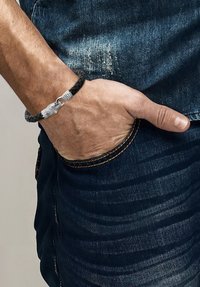 Juodas pynimo odos apyrankė su sidabrine metaline sagtimi, išformuota kaip gyvatė, užsidėta ant riešo, deranti su mėlynais džinsais.
