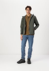 Chaqueta verde con cremallera y capucha, cremallera blanca y muñequera elástica, combinada con una camiseta polo marrón y vaqueros azules. Unos zapatos negros completan el atuendo.