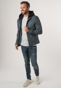 Heren blauwe leren jas met een zwarte capuchon, ritssluiting en geribde manchetten, gecombineerd met versleten jeans en witte sneakers.