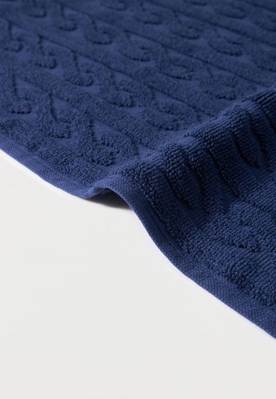 Σκούρο μπλε πετσέτα από πε ρκάλι με ανάγλυφο γεωμετρικό σχέδιο, μερικώς διπλωμένη πάνω σε λευκή επιφάνεια.