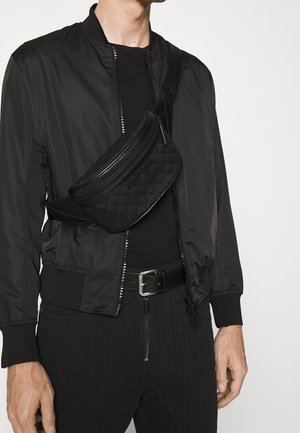 Marsupio nero trapuntato indossato a tracolla, abbinato a una giacca bomber nera e a un top nero aderente. Tessuto strutturato, design elegante.