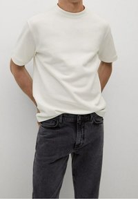 Valkoinen lyhythihainen t-paita pyöreällä pääntiellä, yhdistetty tummanharmaisiin farkkuihin. Kangas näyttää sileältä ja hieman teksturoidulta.