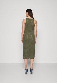 Robe midi en tricot de couleur olive, avec un motif en zigzag texturé, sans manches et avec un ourlet à volants. Portée avec des chaussures à talons hauts bleues.