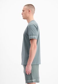 Lichtgrijze t-shirt met korte mouwen en een ronde hals, met het merklogo op de linkermouw. De stof lijkt glad en flexibel.