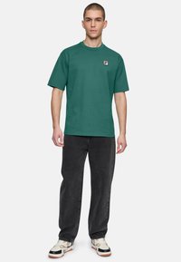 T-shirt verde a maniche corte con scollo rotondo, caratterizzato da un piccolo logo rosso e bianco sul petto. Indossato con jeans neri larghi.