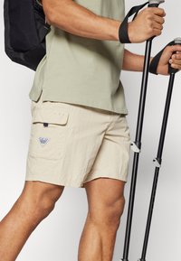 Beige udendørs shorts med et tekstureret stof, der har en sidelomme og et trekantet logo-patch. En sort rygsæk og vandrestave er synlige.