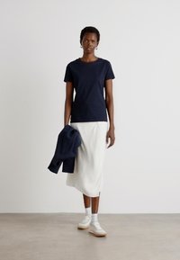 T-shirt blu navy a maniche corte abbinato a una gonna midi bianca, giacca grigia appoggiata sul braccio e sneakers bianche con suola chiara.