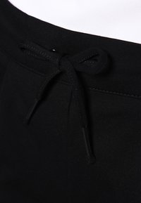 Detailní záběr pásku černých kalhot s prošitými očky a staženou černou šňůrkou na bílém pozadí.