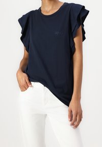 Camiseta azul marino con mangas cortas con volantes y cuello redondo, que presenta un pequeño logotipo de la marca en relieve en el pecho. Combinada con pantalones blancos.