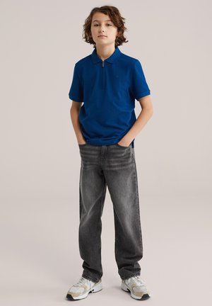 Băiat stând cu mâinile în buzunare, purtând un polo albastru cu fermoar la guler, blugi negri decolorați și adidași albi, pe un fundal simplu.