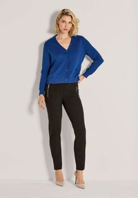 Vrouw die tegen een muur staat, gekleed in een blauwe gestructureerde cardigan met knopen, zwarte broek met ritszakken en beige hoge hakken.