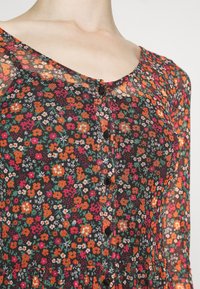 Gros plan sur une robe fleurie transparente avec des boutons noirs et un mélange de fleurs orange, rouges, blanches et vertes sur un fond sombre.