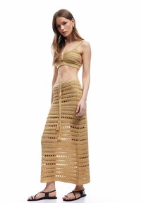 Conjunto de dos piezas de ganchillo en tono marrón claro, que incluye un top de bra de tejido abierto y una falda larga con flecos y agujeros espaciados uniformemente. Sandalias completan el look.