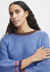 Blauer Strickpullover mit braunem Rippenkragen, strukturiertem Stoff und roten Streifen an den Ärmeln. Das Model posiert mit verschränkten Armen.