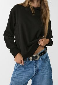 Sweat-shirt noir à coupe décontractée et manches longues, associé à un jean en denim bleu et une ceinture noire avec une boucle argentée décorative.
