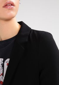 Černý blazer s klopou, hladkým texture a strukturovaným střihem, nošený přes tmavé grafické tričko s výraznými červenými akcenty.