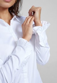 Blusa bianca con colletto classico e polsini a volant. Tessuto liscio con una leggera lucentezza, che mette in risalto una sartoria raffinata e dettagli delicati.