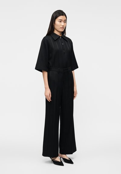 Juodas kombinezonas su trumpomis rankovėmis, sagomis užsegamas priekyje ir plačiomis kelnėmis. Yra vertikalios juostelės ir surišamas diržas. Derinamas su smailiomis balerinkomis.