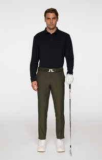 Czarna koszulka polo z długim rękawem, oliwkowe spodnie, biały pasek z detalem logo, białe buty golfowe, trzymający kij golfowy.