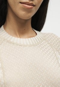 Gros plan d’une personne portant un pull beige à motifs avec un col côtelé rayé, montrant le bas du visage et la zone du cou.