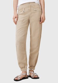 Beige zúžené cargo kalhoty s dvěma předními kapsami, elastickými manžetami a hladkým povrchem. Spárované se stříbrnými sandály na řemínky.