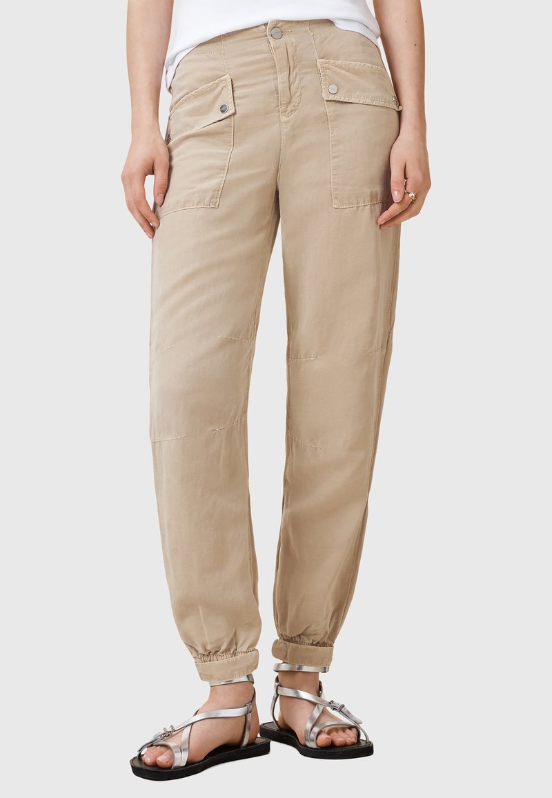 Beige zúžené cargo kalhoty s dvěma předními kapsami, elastickými manžetami a hladkým povrchem. Spárované se stříbrnými sandály na řemínky.