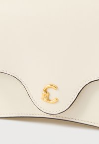 Mala de couro cor creme com um design ondulado e um logótipo dourado em forma de "C". Apresenta costuras pretas contrastantes ao longo das bordas.