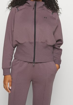 Kvinde iført en mauve jakke med lynlås og matchende bukser med den ene hånd i lommen og krøllet hår synligt.