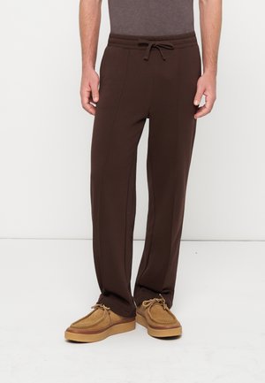 Homme portant un pantalon marron foncé à cordon de serrage et des chaussures en daim beige à lacets, debout devant un fond blanc uni.