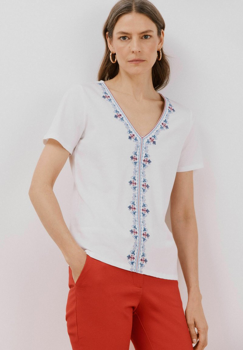 Haut blanc à manches courtes avec un col en V et une broderie florale bleue et rouge au centre. Assorti à un pantalon rouge. Texture lisse.