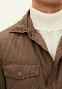 Giubbotto imbottito marrone con due tasche sul petto, caratterizzato da una chiusura con bottoni e un maglione bianco a coste con colletto alto sotto. Texture liscia.