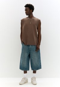 Camicetta marrone senza maniche, vestibilità rilassata, abbinata a pantaloni corti in denim leggero blu chiaro. Indossata con calzini bianchi e sneakers beige. Collane argentate a strati.