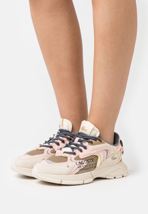 Jambes portant des baskets Lacoste roses, beige et grises avec des panneaux en mesh, des accents en daim et des lacets noirs, posées sur une surface blanche.