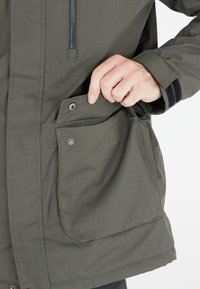 Giacca da esterno verde con chiusura a zip, grande tasca con patta e tessuto testurizzato. I polsini presentano dettagli in Velcro neri.
